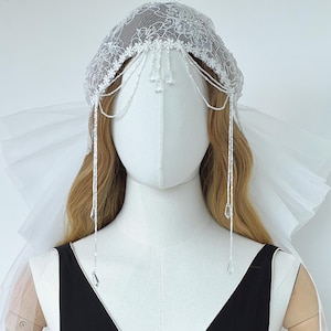 Könnte beinhalten: Ein weißer Kopfschmuck aus Spitze mit filigraner Blumenstickerei und zarten Perlenverzierungen. Das Accessoire umfasst drapierte Perlenstränge mit klaren, tropfenförmigen Kristallanhängern, die über die Stirn fallen, sowie einen weißen Tüllschleier.