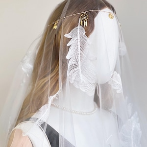 May include: A white bridal veil with embroidered feather details and a gold-toned headband. The headband features small charms and a dreamcatcher pendant. The veil is draped over a mannequin, showcasing the delicate design and texture of the fabric.
