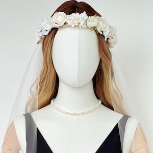 May include: A floral crown featuring white and cream-colored fabric flowers and green leaves, paired with a sheer white veil. The accessory is displayed on a mannequin wearing a simple pearl necklace and a black sleeveless garment with a V-neckline.