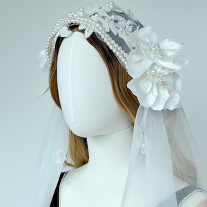 Peut inclure: Un accessoire de tête de mariée blanc composé d'un voile transparent orné de petites fleurs en tissu et de perles. Le serre-tête est décoré de dentelle blanche, de rangées de perles et de grandes fleurs en tissu blanc sur les côtés.