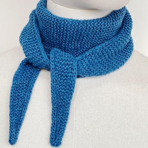 Könnte beinhalten: Ein leuchtend blaugrüner Strickschal mit einer strukturierten kraus rechts gestrickten Oberfläche. Der Schal ist um den Hals einer Schaufensterpuppe gewickelt, wobei zwei spitz zulaufende Enden vorne herabhängen und eine gleichmäßige Strickstruktur aufweisen.