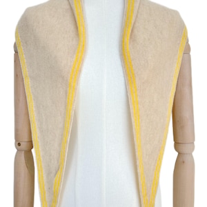 May include: A triangular scarf in a soft beige knit fabric featuring bright yellow stripes along the edges. The scarf is draped around the neck, showcasing a pointed design that tapers downward, with the yellow detailing framing the entire perimeter.