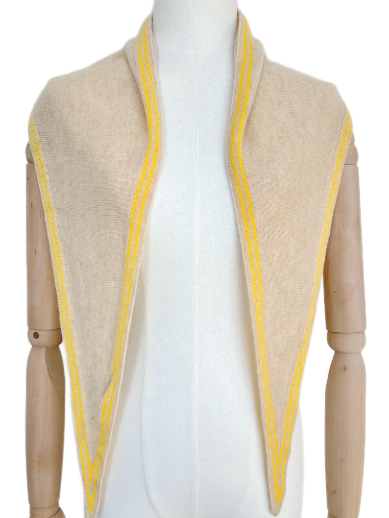 May include: A triangular scarf in a soft beige knit fabric featuring bright yellow stripes along the edges. The scarf is draped around the neck, showcasing a pointed design that tapers downward, with the yellow detailing framing the entire perimeter.