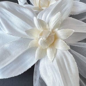 May include: Close-up of white fabric flower hair accessories. The flowers have multiple petals and a creamy center. The accessories are made of a soft, silky material and are attached to a fine mesh netting.