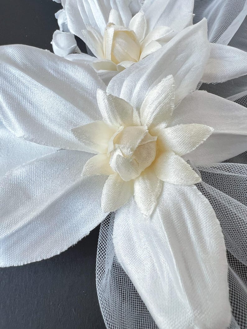 May include: Close-up of white fabric flower hair accessories. The flowers have multiple petals and a creamy center. The accessories are made of a soft, silky material and are attached to a fine mesh netting.