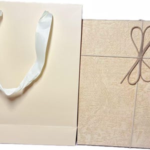 Op de afbeelding: Een lichtbeige papieren cadeauzak met witte linthengsels naast een rechthoekige cadeauverpakking met een getextureerd beige oppervlak en een decoratief bruin elastisch koord dat als strik op de voorzijde is gebonden.