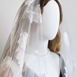 May include: A white tulle veil with embroidered feather details. The veil is draped over a mannequin head with brown hair and a pearl necklace. The veil's delicate design adds a touch of elegance.
