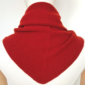 Könnte beinhalten: Ein einfarbiger, roter, dreieckiger Strickschal, der um den Hals einer weißen Schaufensterpuppe drapiert ist. Der Schal weist eine feine, gleichmäßige Rippenstruktur auf und hat eine spitz zulaufende Vorderkante, die eine geschichtete Optik erzeugt.