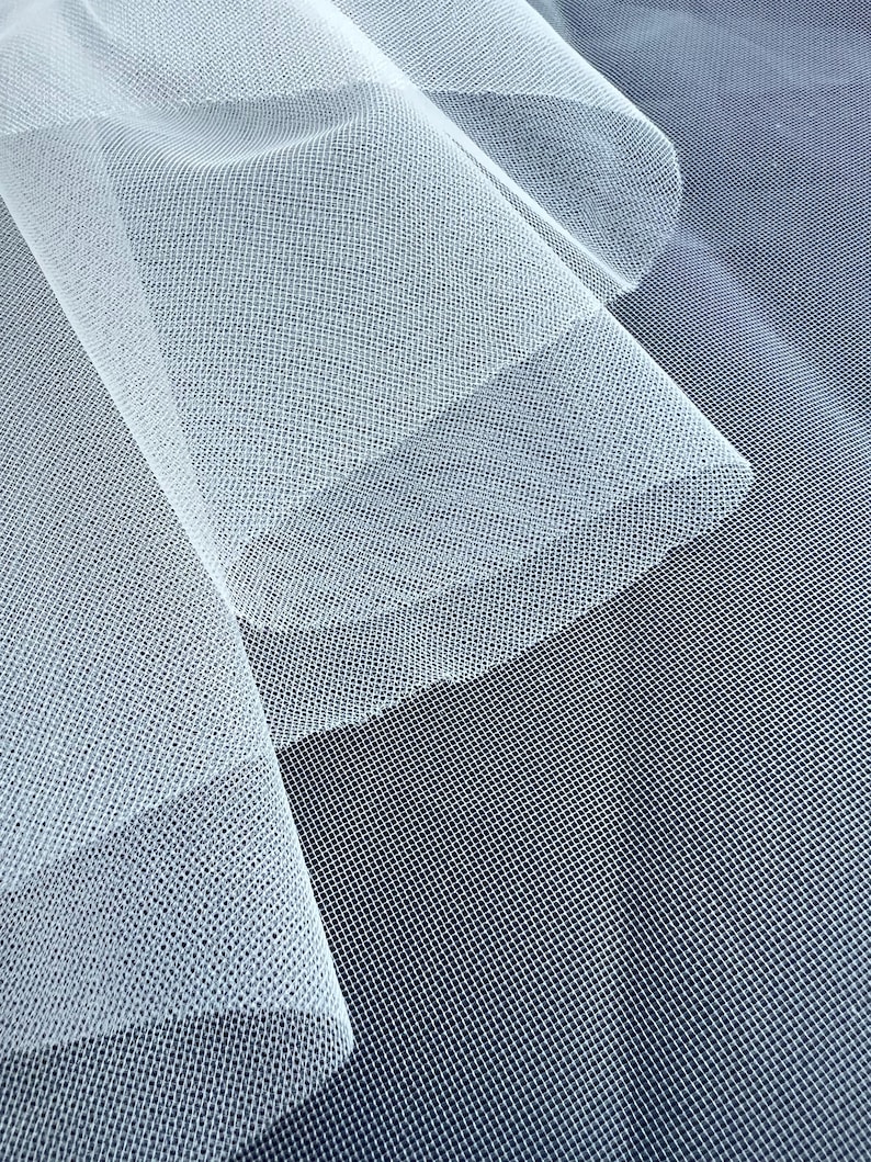 May include: Close-up of white tulle fabric with a fine mesh texture. The fabric is layered, creating depth and shadows. The background is a darker shade of blue, providing contrast to the delicate, sheer material.