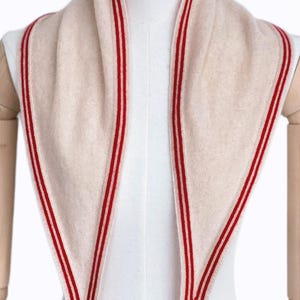 May include: A triangular scarf in a soft beige color featuring a distinct red and white striped border along the edges. The knit fabric has a smooth texture and is draped symmetrically around the neck area, creating two long pointed ends in front.