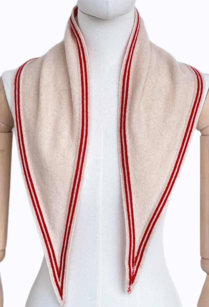 May include: A triangular scarf in a soft beige color featuring a distinct red and white striped border along the edges. The knit fabric has a smooth texture and is draped symmetrically around the neck area, creating two long pointed ends in front.
