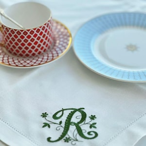 Könnte beinhalten: Eine weiße Leinen-Serviette mit einem grünen floralen "R" wird mit einer rot-weiß gemusterten Tasse und Untertasse sowie einem hellblauen Teller mit Goldrand präsentiert. In der Tasse befindet sich ein goldener Löffel.