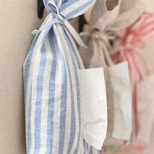 May include: A blue and white striped linen tissue holder hangs from a black hook. The holder is tied at the top with a matching fabric tie. Other tissue holders in neutral colors are visible in the background.