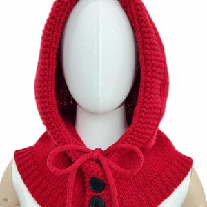 Könnte beinhalten: Eine leuchtend rote Strickhaube mit passendem Kragen. Die Kapuze hat eine abgerundete Form und ist mit einem Kragen verbunden, der mit zwei schwarzen Knöpfen und einer Krawatte befestigt wird. Die Strickstruktur ist im gesamten Design sichtbar.