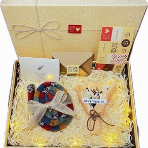 May include: A gift box with a beige lid and a bow, filled with decorative straw, a colorful knitted item, a small bouquet, stickers, and cards with the text "Just for you" and "Best Wishes".