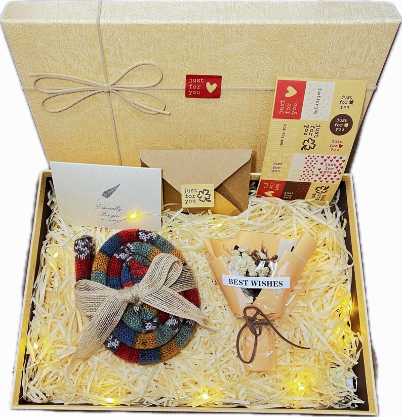 May include: A gift box with a beige lid and a bow, filled with decorative straw, a colorful knitted item, a small bouquet, stickers, and cards with the text "Just for you" and "Best Wishes".