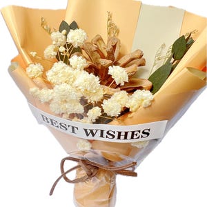 May include: A bouquet of dried flowers and a pinecone wrapped in tan paper. The bouquet includes white flowers and greenery, tied with a brown ribbon. The words "BEST WISHES" are printed on a white band around the bouquet.