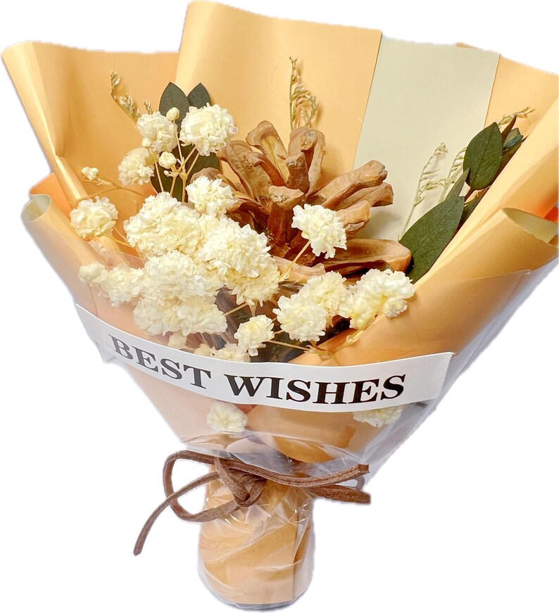 May include: A bouquet of dried flowers and a pinecone wrapped in tan paper. The bouquet includes white flowers and greenery, tied with a brown ribbon. The words "BEST WISHES" are printed on a white band around the bouquet.