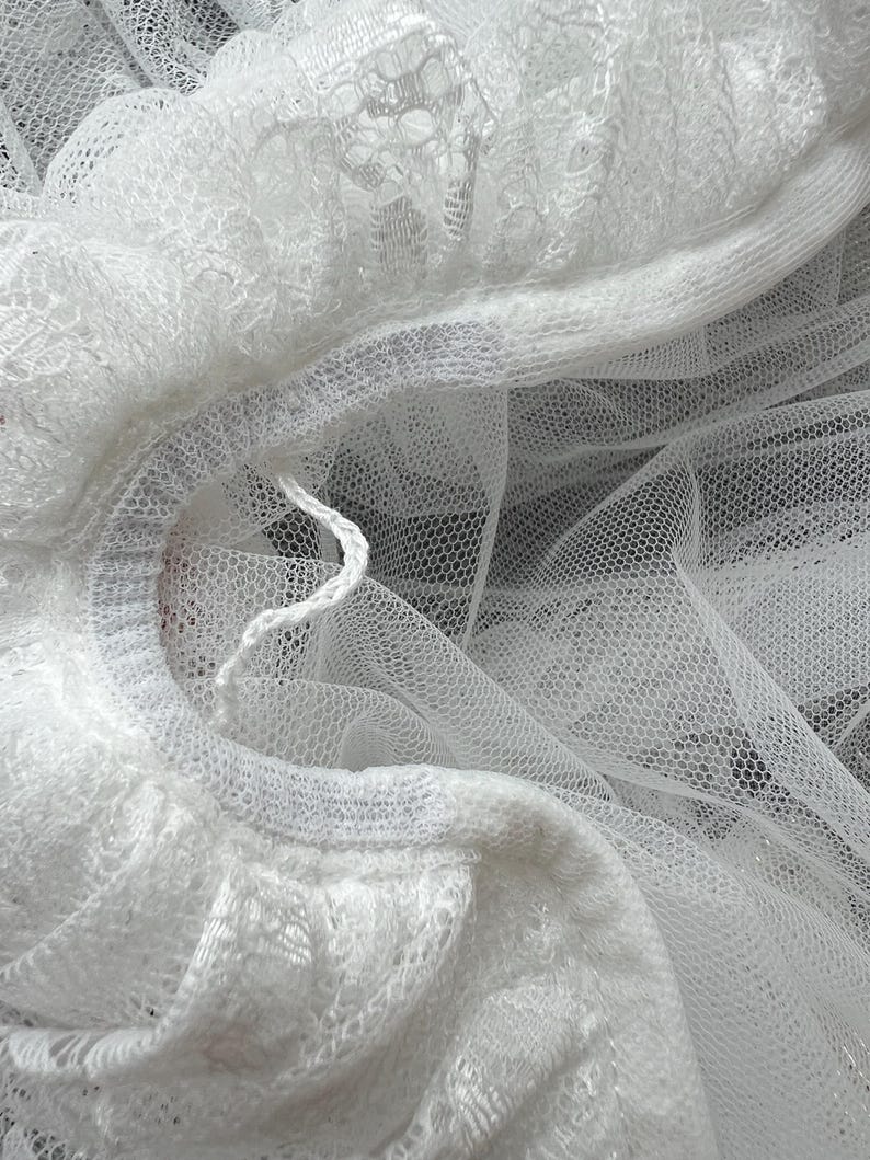 Op de afbeelding: Een witte tule stof met een delicate bloemenkantrand langs de rand. Het materiaal is doorschijnend en gelaagd, met een fijne gaasstructuur en een enkele losse witte draad zichtbaar bij het geplooide, kanten randdetail.