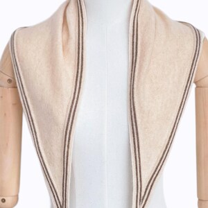 May include: A triangular scarf in a soft beige hue, featuring a fine, dark brown striped border along the edges. The scarf is draped around the neck of a mannequin, showcasing a smooth, knit texture and a pointed, symmetrical design.