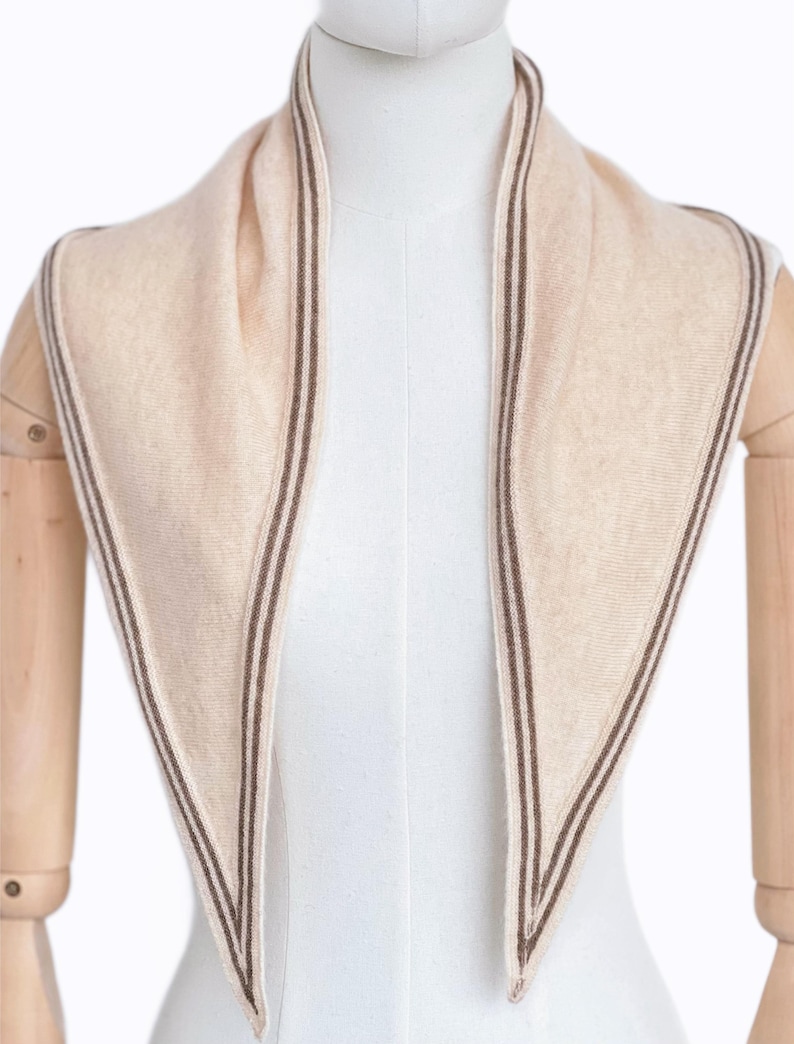 May include: A triangular scarf in a soft beige hue, featuring a fine, dark brown striped border along the edges. The scarf is draped around the neck of a mannequin, showcasing a smooth, knit texture and a pointed, symmetrical design.