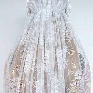 Op de afbeelding: Een witte kanten sluier met een gedetailleerd bloemmotief over de gehele stof. De sluier is aan de bovenkant geplooid en aan beide zijden versierd met witte stoffen bloemen, die in lange, doorschijnende en semi-transparante lagen naar beneden vallen.