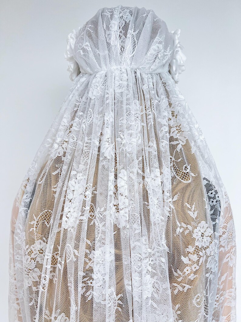 Op de afbeelding: Een witte kanten sluier met een gedetailleerd bloemmotief over de gehele stof. De sluier is aan de bovenkant geplooid en aan beide zijden versierd met witte stoffen bloemen, die in lange, doorschijnende en semi-transparante lagen naar beneden vallen.