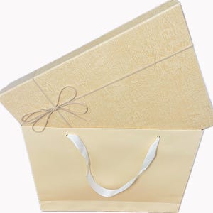 May include: A cream-colored gift box with a textured surface and a matching shopping bag. The box is tied with a thin, light brown string and a bow. The shopping bag has white ribbon handles.