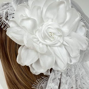 Op de afbeelding: Een grote witte satijnen stoffen bloem met gelaagde bloemblaadjes, bevestigd aan een witte kanten sluier met een geschulpte rand. Het bloemstuk is gecentreerd op het net, wat zorgt voor een gelaagde uitstraling met gladde, glanzende stof.