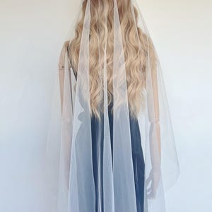 May include: A bridal veil made of sheer white fabric, draped over a mannequin. The veil cascades down, partially obscuring the mannequin's back and a black dress. The mannequin has long, wavy blonde hair and a floral headpiece.