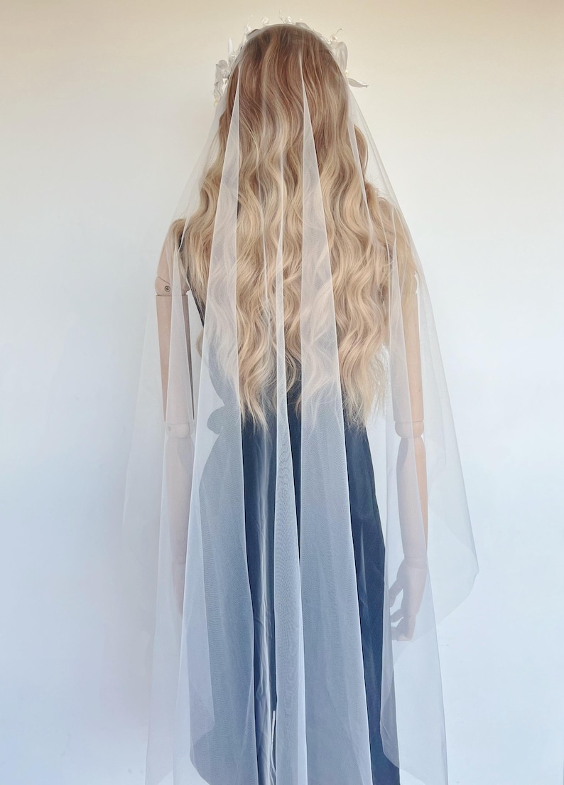 May include: A bridal veil made of sheer white fabric, draped over a mannequin. The veil cascades down, partially obscuring the mannequin's back and a black dress. The mannequin has long, wavy blonde hair and a floral headpiece.
