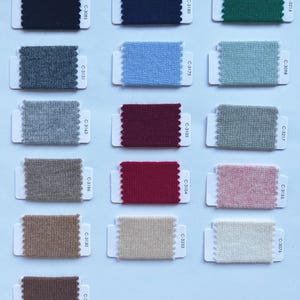 May include: A collection of sixteen rectangular fabric swatches arranged in a grid, each attached to a white card with a scalloped edge. The swatches feature various solid colors including black, navy, green, gray, blue, red, pink, beige, and brown.