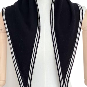 May include: A black triangular scarf featuring a white double-striped border along the edges. The knit fabric is solid black with clean, parallel white lines running along the perimeter, creating a sharp contrast against the dark background.