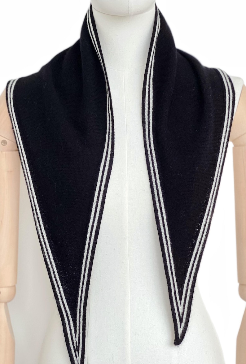 May include: A black triangular scarf featuring a white double-striped border along the edges. The knit fabric is solid black with clean, parallel white lines running along the perimeter, creating a sharp contrast against the dark background.