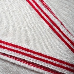 May include: A cream-colored knit fabric featuring prominent red stripes along the edges. The material displays a fine, consistent texture with a soft appearance, showcasing the detailed weave of the textile and the bold contrast of the red lines.