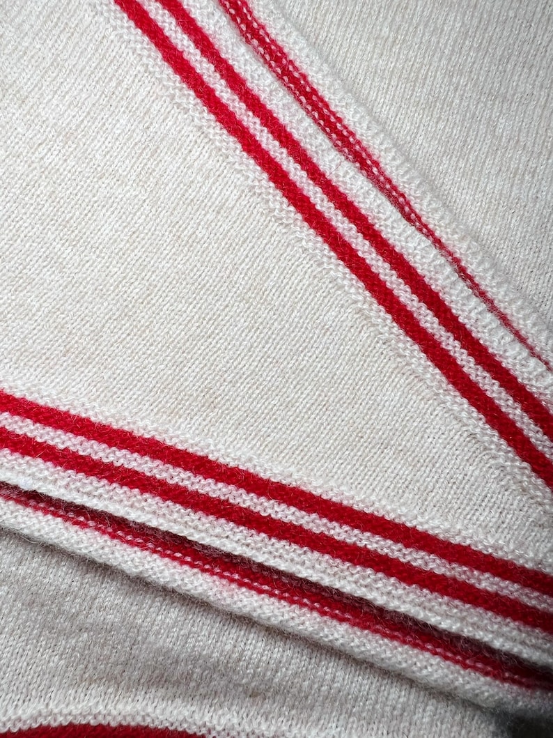 May include: A cream-colored knit fabric featuring prominent red stripes along the edges. The material displays a fine, consistent texture with a soft appearance, showcasing the detailed weave of the textile and the bold contrast of the red lines.