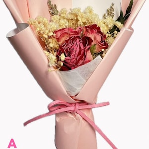 May include: A bouquet of dried red roses and small white filler flowers wrapped in light pink paper. The arrangement is secured with a pink suede-like ribbon tied in a bow. The letter A is printed in pink in the bottom left corner of the image.