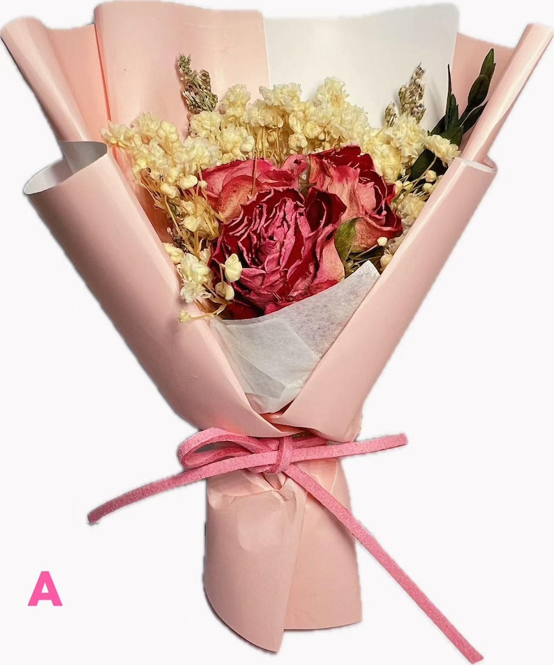 May include: A bouquet of dried red roses and small white filler flowers wrapped in light pink paper. The arrangement is secured with a pink suede-like ribbon tied in a bow. The letter A is printed in pink in the bottom left corner of the image.