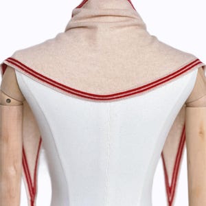 May include: A beige knit triangular scarf featuring a prominent red striped border along the edges. The scarf is draped around the neck area, with two pointed ends extending downwards, showcasing a consistent red and beige color scheme throughout.