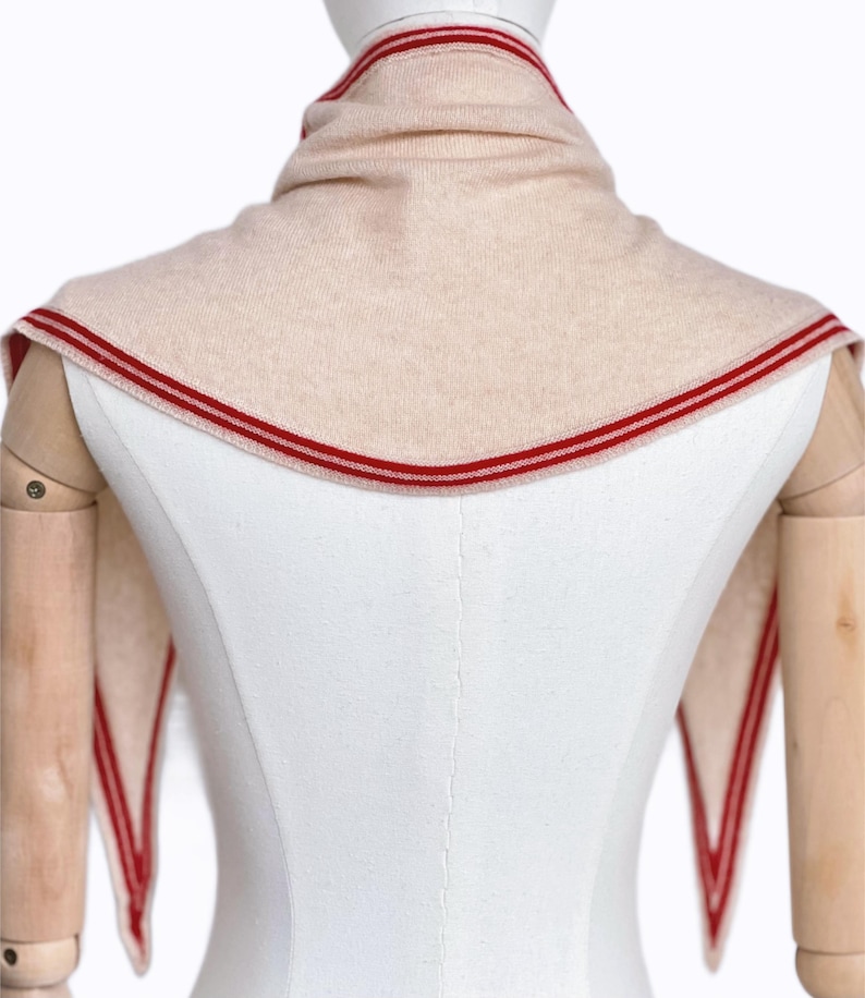 May include: A beige knit triangular scarf featuring a prominent red striped border along the edges. The scarf is draped around the neck area, with two pointed ends extending downwards, showcasing a consistent red and beige color scheme throughout.