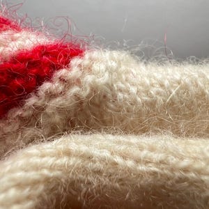 May include: A close-up view of a knitted textile featuring a fuzzy, brushed texture. The fabric displays alternating horizontal stripes of cream and vibrant red yarn, with loose, fine fibers creating a soft, halo-like appearance across the surface.