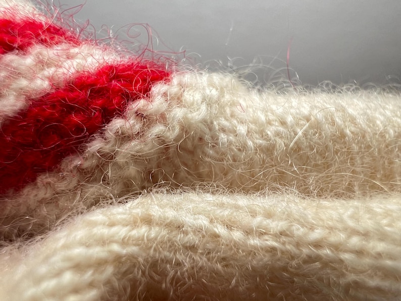 May include: A close-up view of a knitted textile featuring a fuzzy, brushed texture. The fabric displays alternating horizontal stripes of cream and vibrant red yarn, with loose, fine fibers creating a soft, halo-like appearance across the surface.