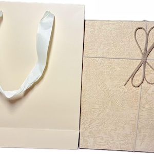 May include: A light beige paper gift bag with white ribbon handles positioned next to a rectangular gift box featuring a textured tan surface and a decorative brown cord tied in a bow across the front of the lid.