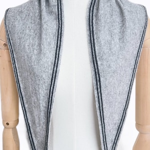 May include: A light gray triangular scarf featuring a textured knit construction. The edges of the scarf are detailed with two thin, parallel dark charcoal stripes that run along the entire perimeter of the fabric, creating a clean, linear border.