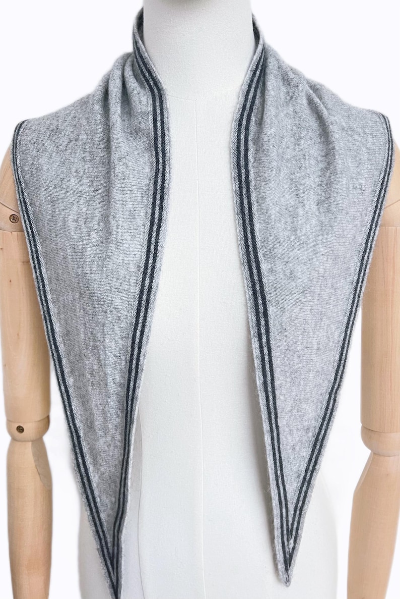 May include: A light gray triangular scarf featuring a textured knit construction. The edges of the scarf are detailed with two thin, parallel dark charcoal stripes that run along the entire perimeter of the fabric, creating a clean, linear border.