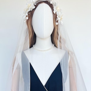 May include: A white bridal headpiece with floral accents and a sheer veil. The headpiece features delicate white flowers and pearl-like beads. The veil is draped over a mannequin wearing a navy blue dress and a pearl necklace.