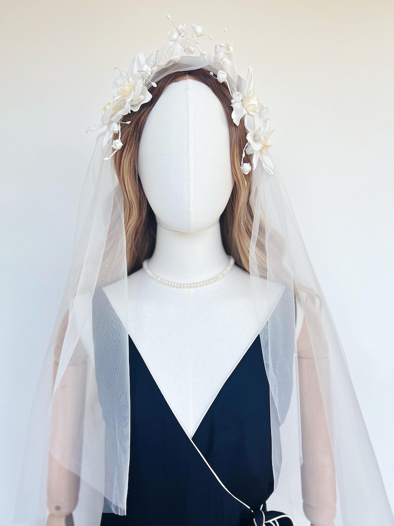 May include: A white bridal headpiece with floral accents and a sheer veil. The headpiece features delicate white flowers and pearl-like beads. The veil is draped over a mannequin wearing a navy blue dress and a pearl necklace.