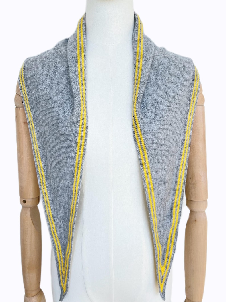 May include: A triangular scarf in heather gray knit fabric featuring two bright yellow stripes running parallel along each of the three edges. The scarf is draped over a mannequin, displaying a textured surface and pointed ends.