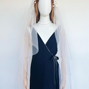 May include: A navy blue wrap dress with a white trim and a matching bow. The dress is paired with a sheer white veil and a floral crown. A pearl necklace completes the bridal look.
