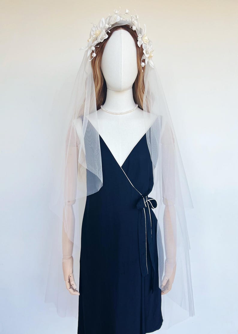 May include: A navy blue wrap dress with a white trim and a matching bow. The dress is paired with a sheer white veil and a floral crown. A pearl necklace completes the bridal look.